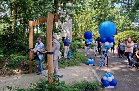 Ceremonie met lintdoorknippen bij houten boog, omgeven door ballonnen, publiek en fotografen.