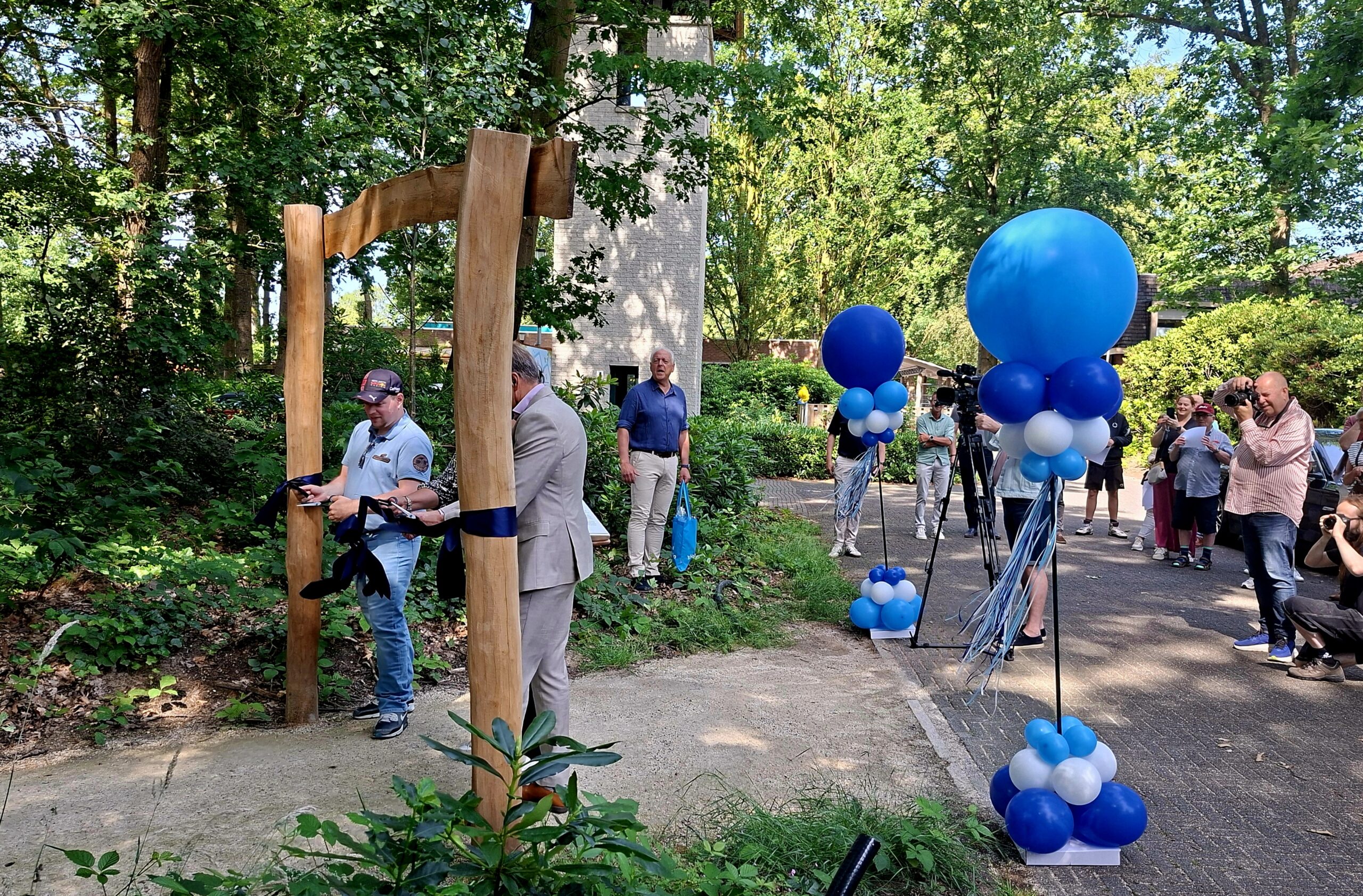 Ceremonie met lintdoorknippen bij houten boog, omgeven door ballonnen, publiek en fotografen.