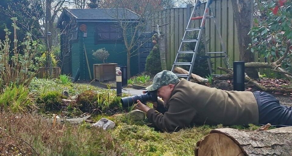 Persoon met cameralens ligt plat in tuin, richt zich op iets. Achtergrond: schuurtje en ladder.
