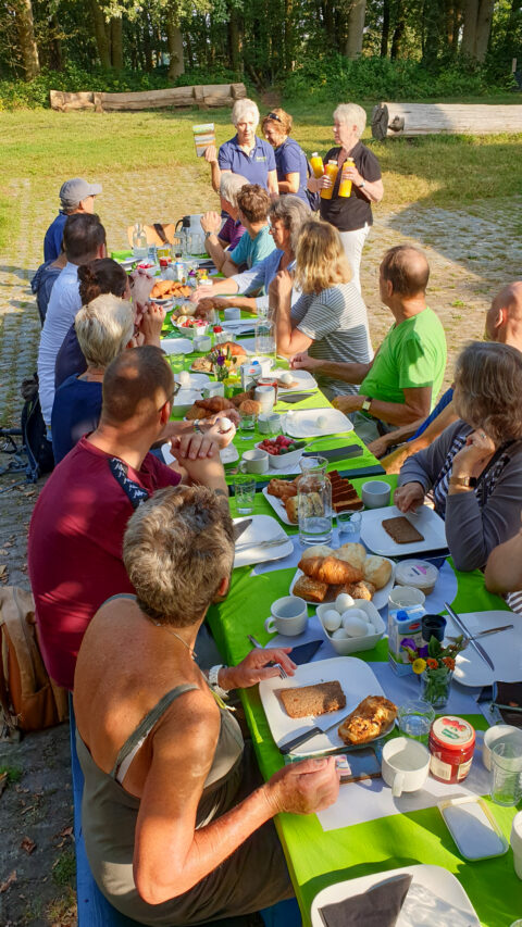 Groepsontbijt buiten aan lange tafel met diverse gerechten, mensen luisteren naar staande spreker.