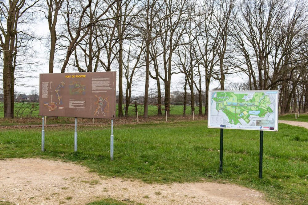 Informatieborden bij Fort de Roovere, met wandelkaart en tekst, omgeven door bomen en grasveld.
