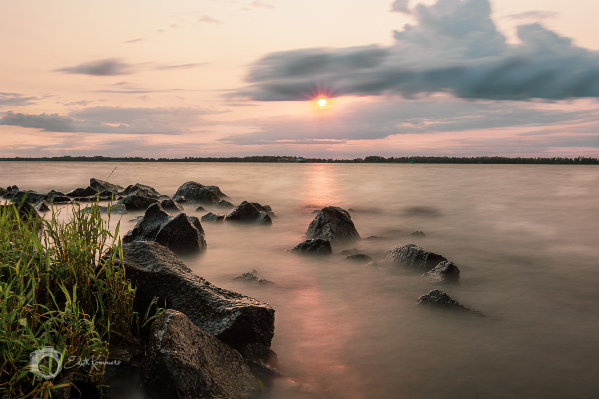 Rotsachtige oever bij zonsondergang, met rustige golven en grassen onder een bewolkte hemel.