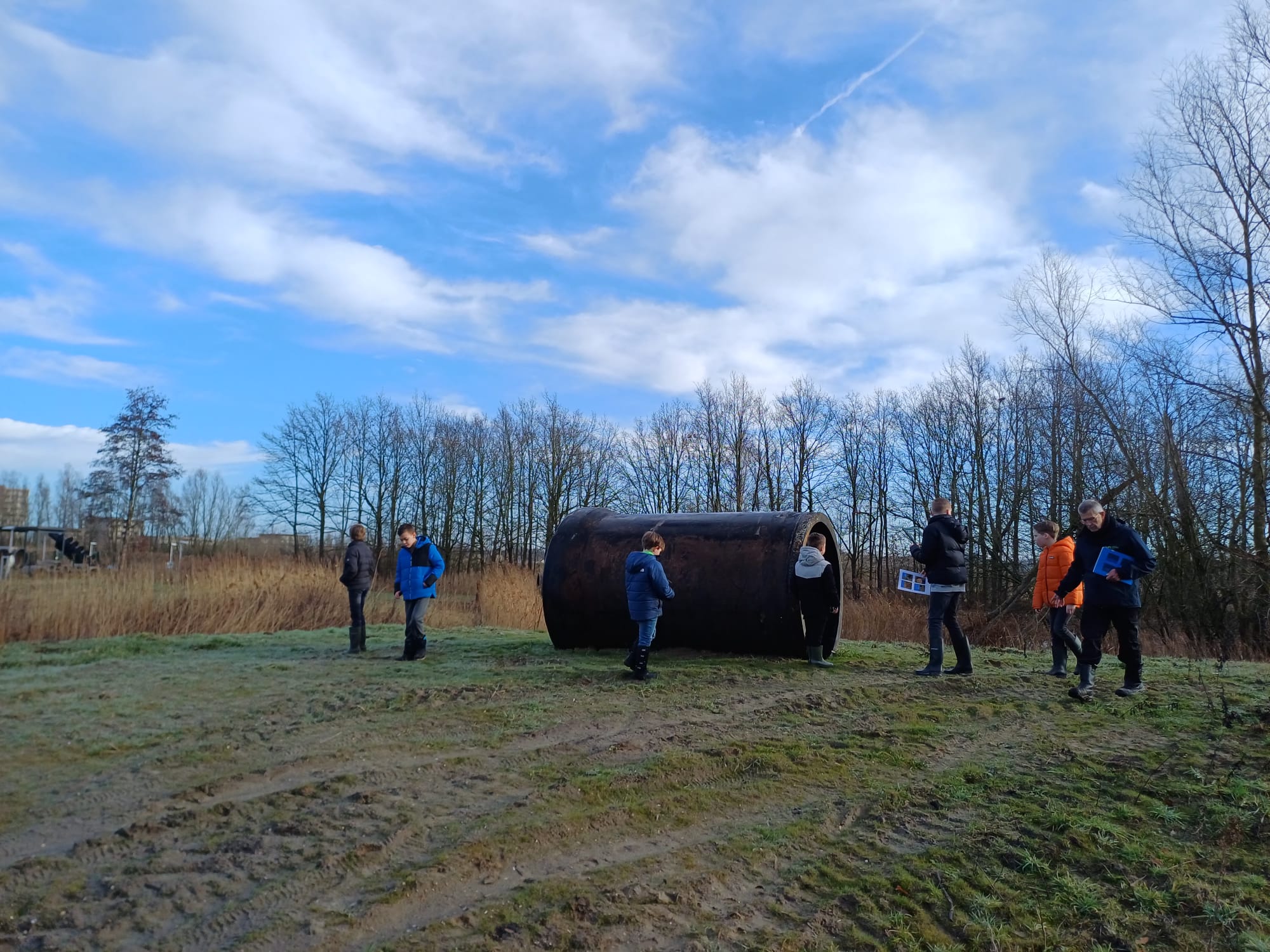 Groep mensen loopt buiten bij een groot, cilindrisch kunstobject op een grasveld met bomen op de achtergrond.