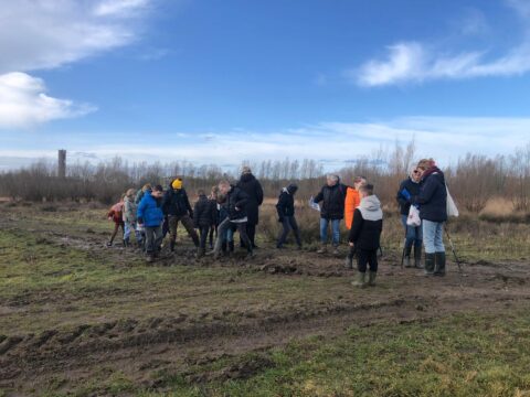 Een groep mensen wandelt in een modderig veld onder een blauwe lucht met wolken.