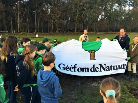 Kinderen in scoutsuniform houden een spandoek vast met de tekst "Geef om de Natuur" in een bosgebied.