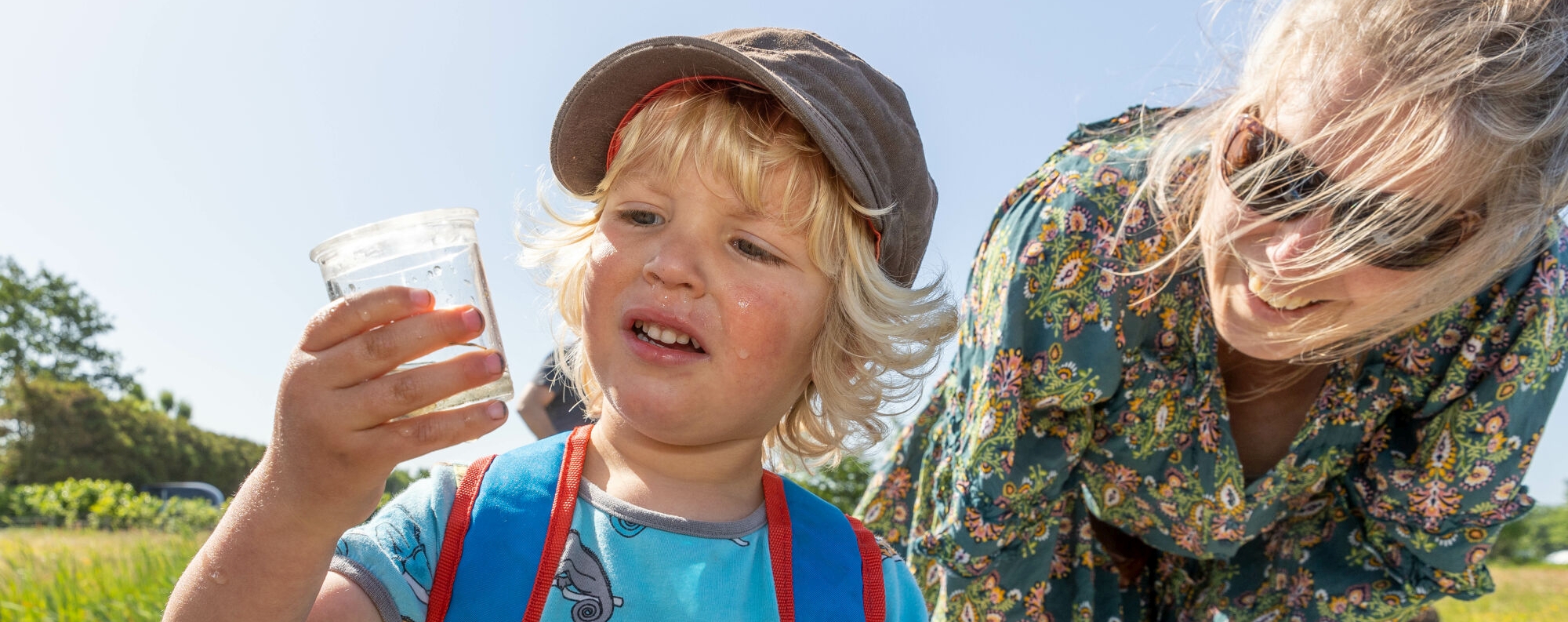 Een kind kijkt nieuwsgierig naar een plastic beker buiten, begeleid door een lachende volwassene.