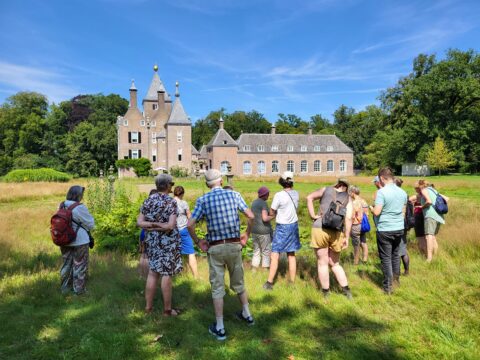 Groep mensen kijkt naar een historisch kasteel omringd door groen onder een blauwe hemel.