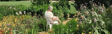 Vrouw zit op witte stoel in bloementuin, genietend van groene natuur en kleurrijke bloemen.