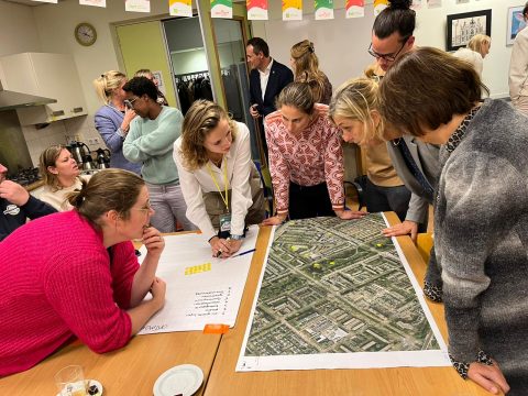 Groep mensen buigt zich over een grote plattegrond op tafel in een vergaderruimte.