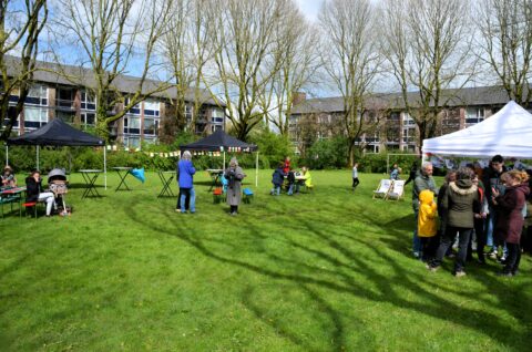 Buitenbijeenkomst op een grasveld met mensen rond tenten, omgeven door bomen en flats.
