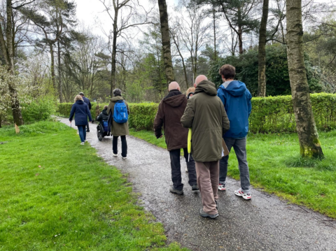Groep mensen wandelt op een pad in een bosrijke omgeving, waaronder iemand in een rolstoel.
