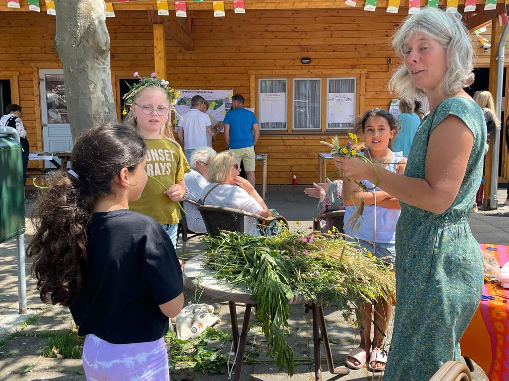 Vrouwen en kinderen maken bloemenkransen bij een houten gebouw op een zonnige dag.