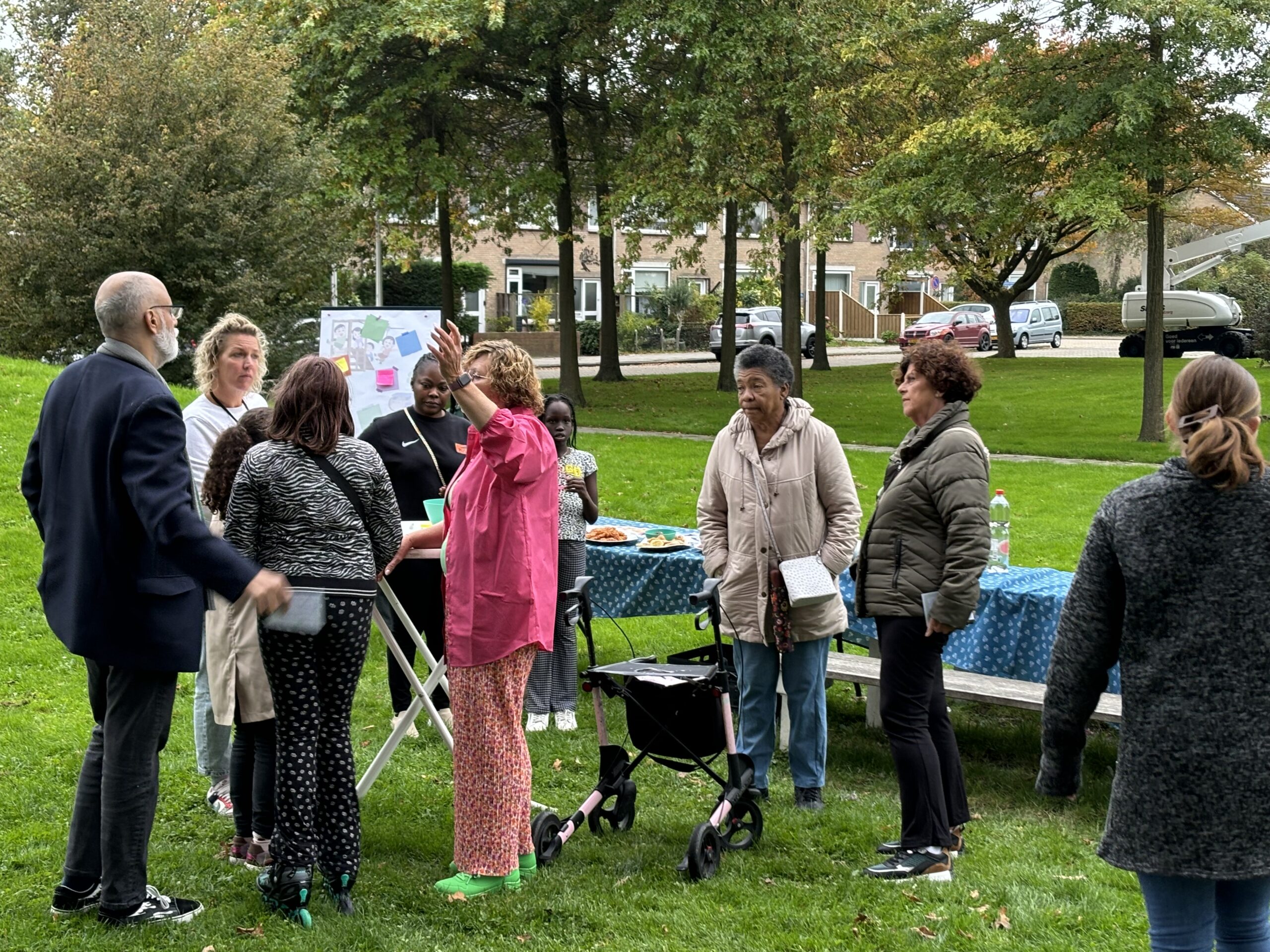 Groep mensen in gesprek in een park, bij een tafel met snacks en een bord met notities.