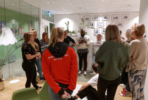Groep mensen in een kring, praten in een kantooromgeving met planten en fotolijstjes op de muur.