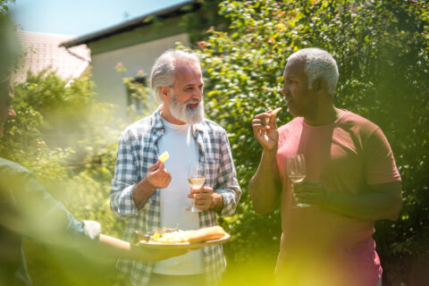 Twee mannen praten en snacken in een tuin, met een dienblad en wijnglazen in hun handen.