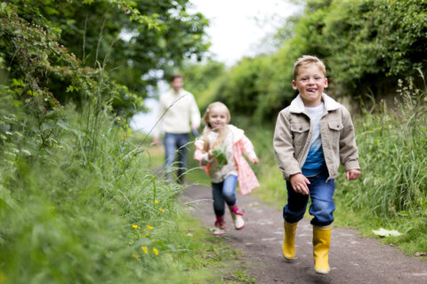 Kinderen rennen lachend over een bospaadje, omringd door groen en bloemen. Volwassene op de achtergrond.
