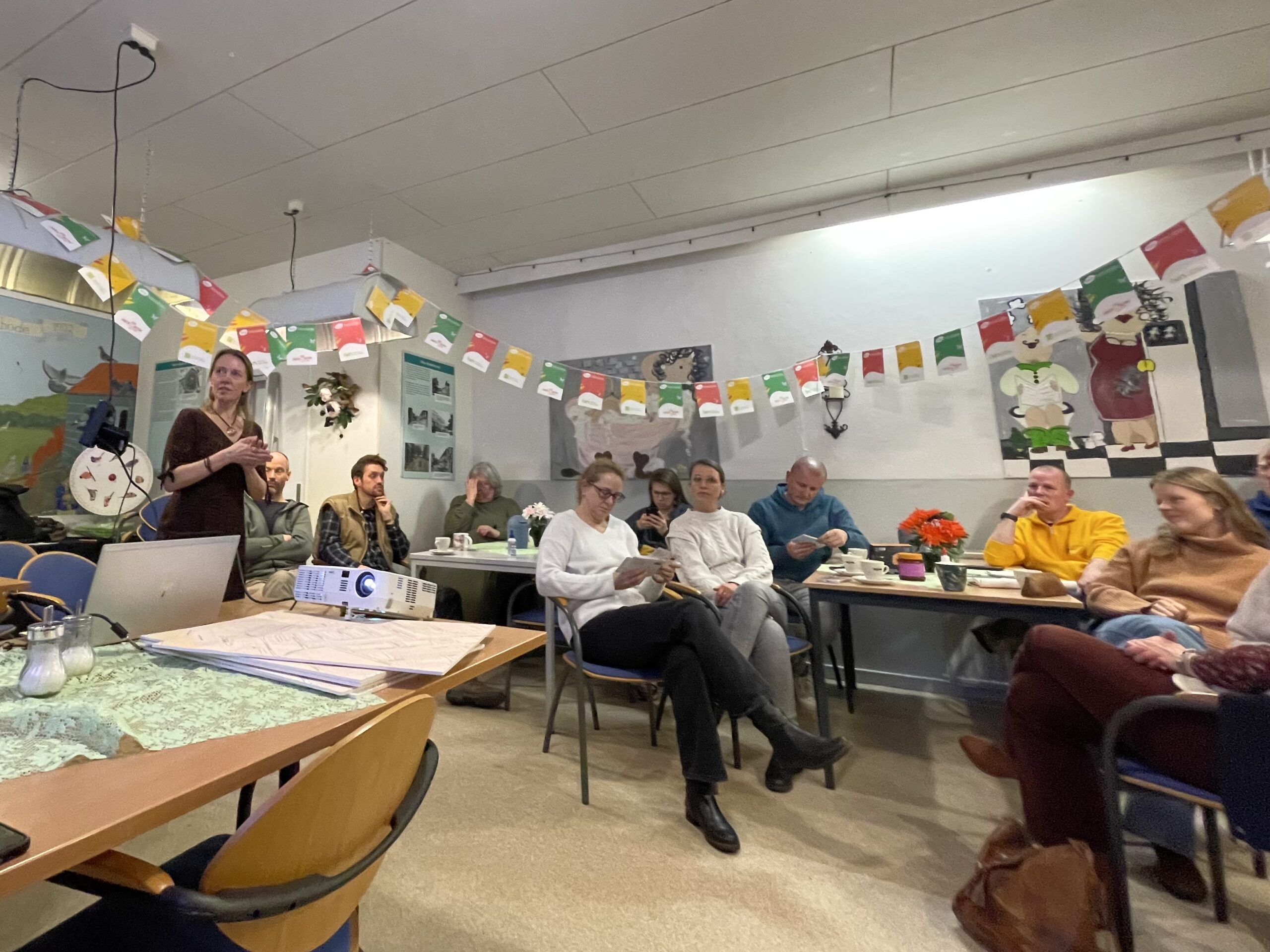 Mensen in een kamer luisteren naar een presentatie, met kleurrijke slingers en een projector op tafel.