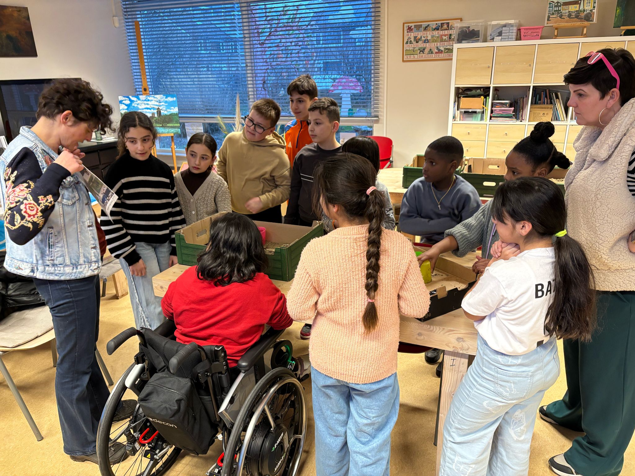 Kinderen en volwassenen staan rond een tafel in een klaslokaal, iemand in een rolstoel.