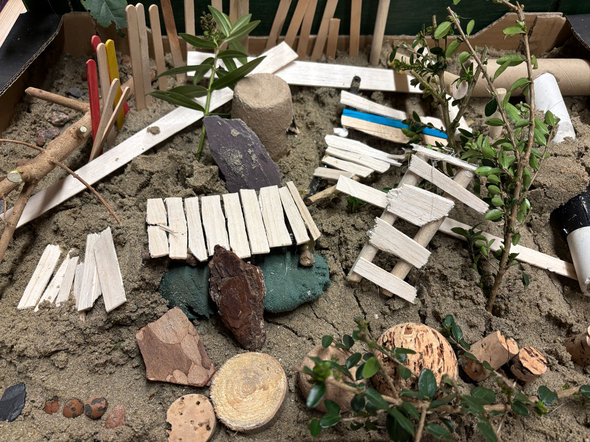 Miniatuurtuin met zand, houten hekjes, takken en kleine plantjes.