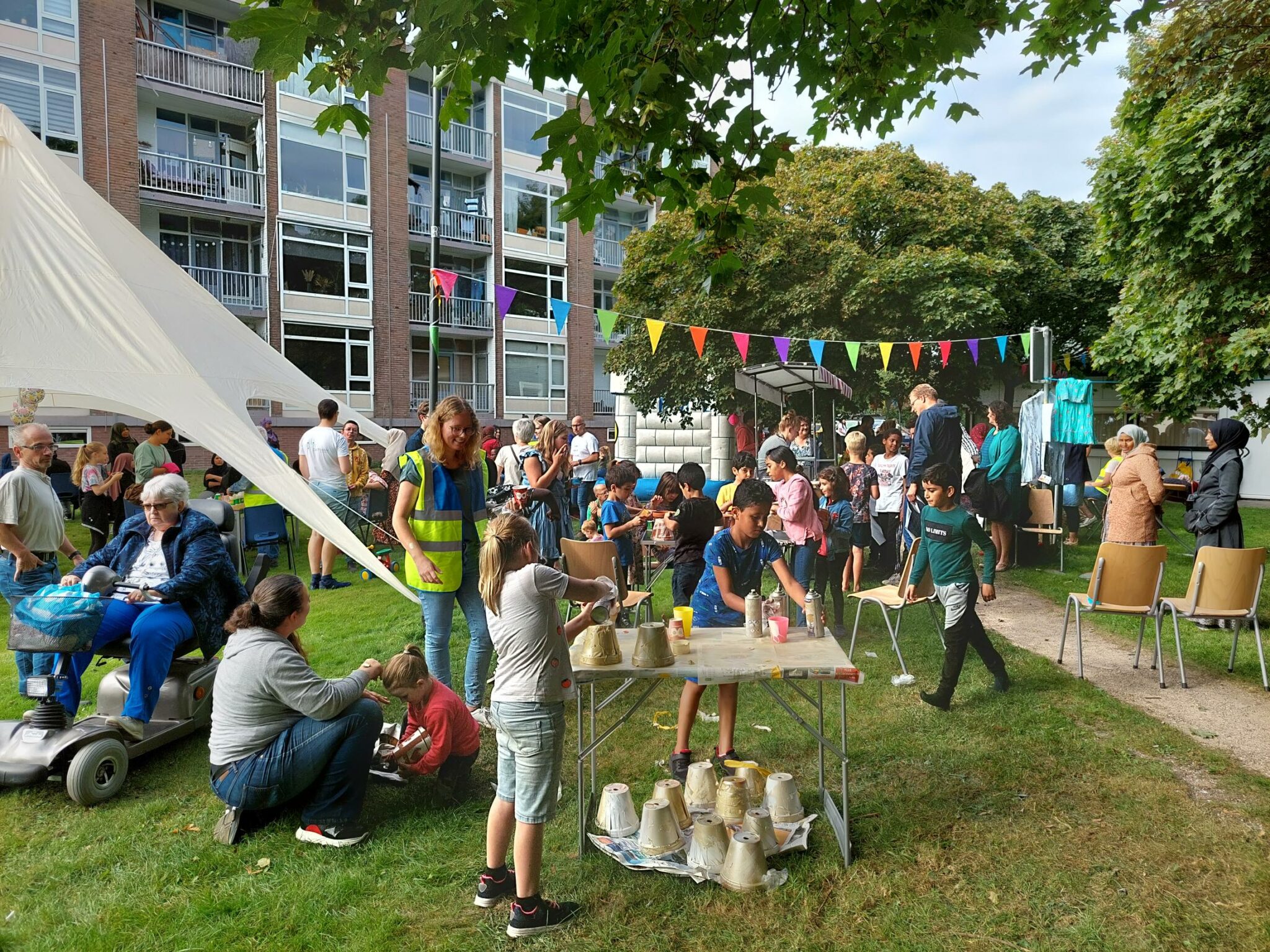 Buitenfeest met mensen onder kleurrijke vlaggen nabij een gebouw; kinderen verven potten.
