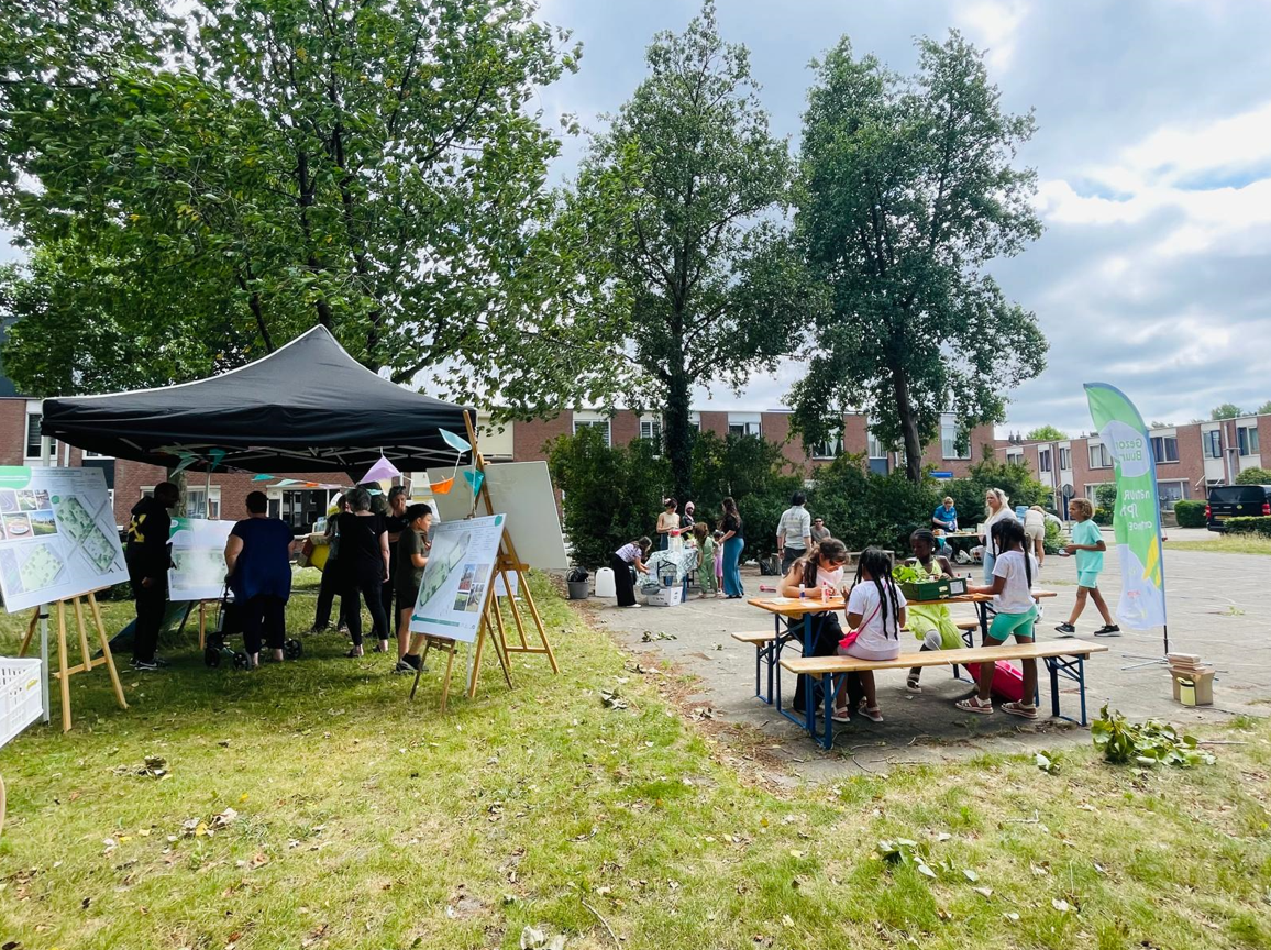Buitenevenement met tent, posters en kinderen aan tafels op een grasveld, omgeven door bomen en huizen.