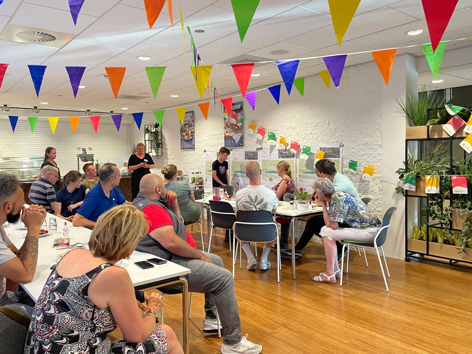 Groep mensen zit in een versierde ruimte met gekleurde vlaggetjes, aandachtig luisterend naar een spreker.