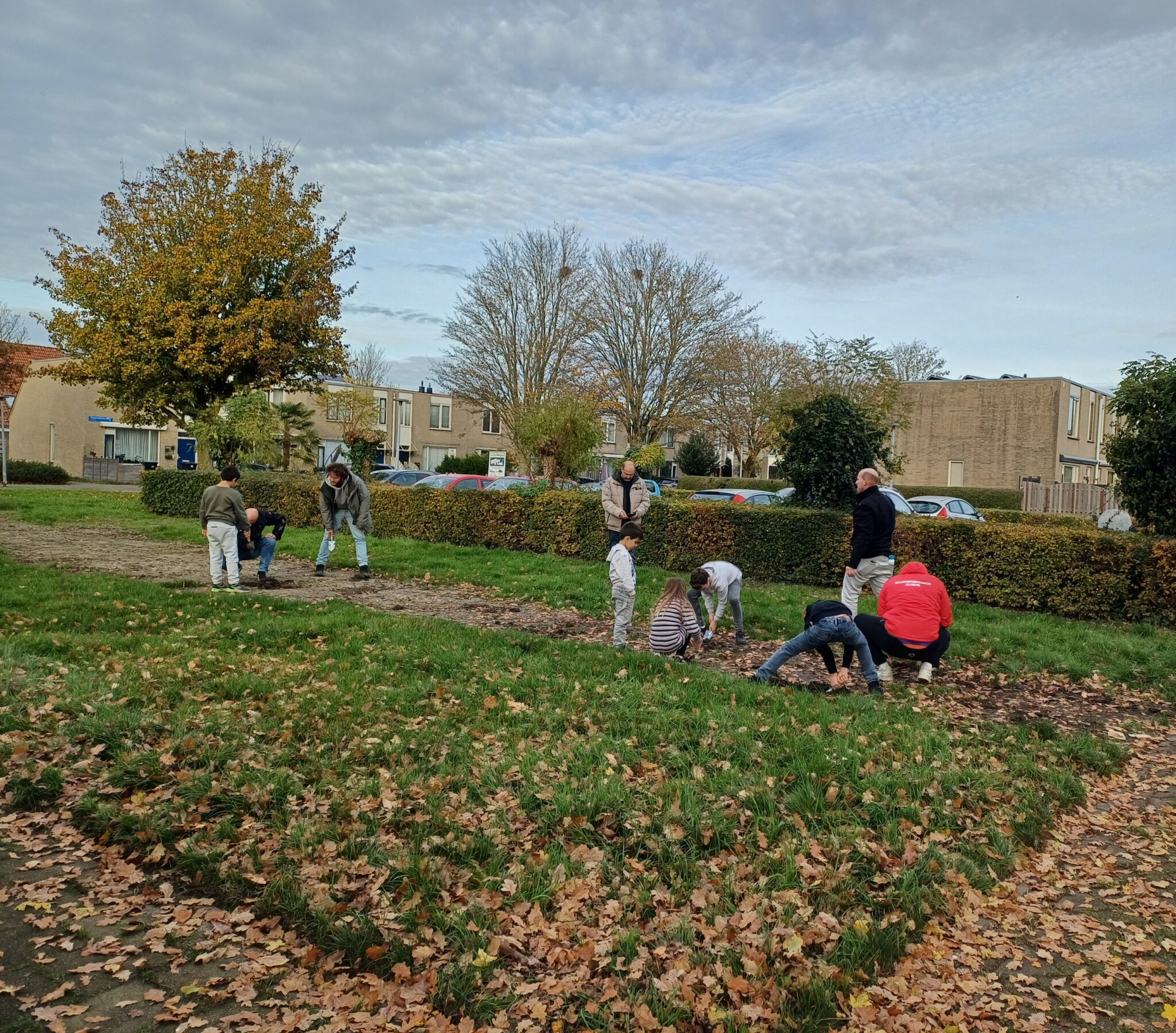 Mensen planten bollen in een herfstig park, omringd door huizen en kale bomen.