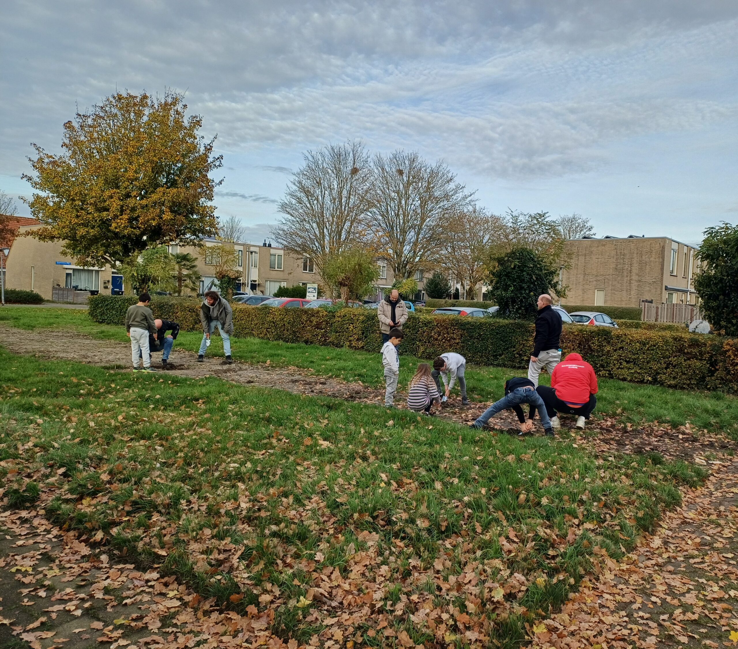 Mensen planten bollen in een herfstig park, omringd door huizen en kale bomen.