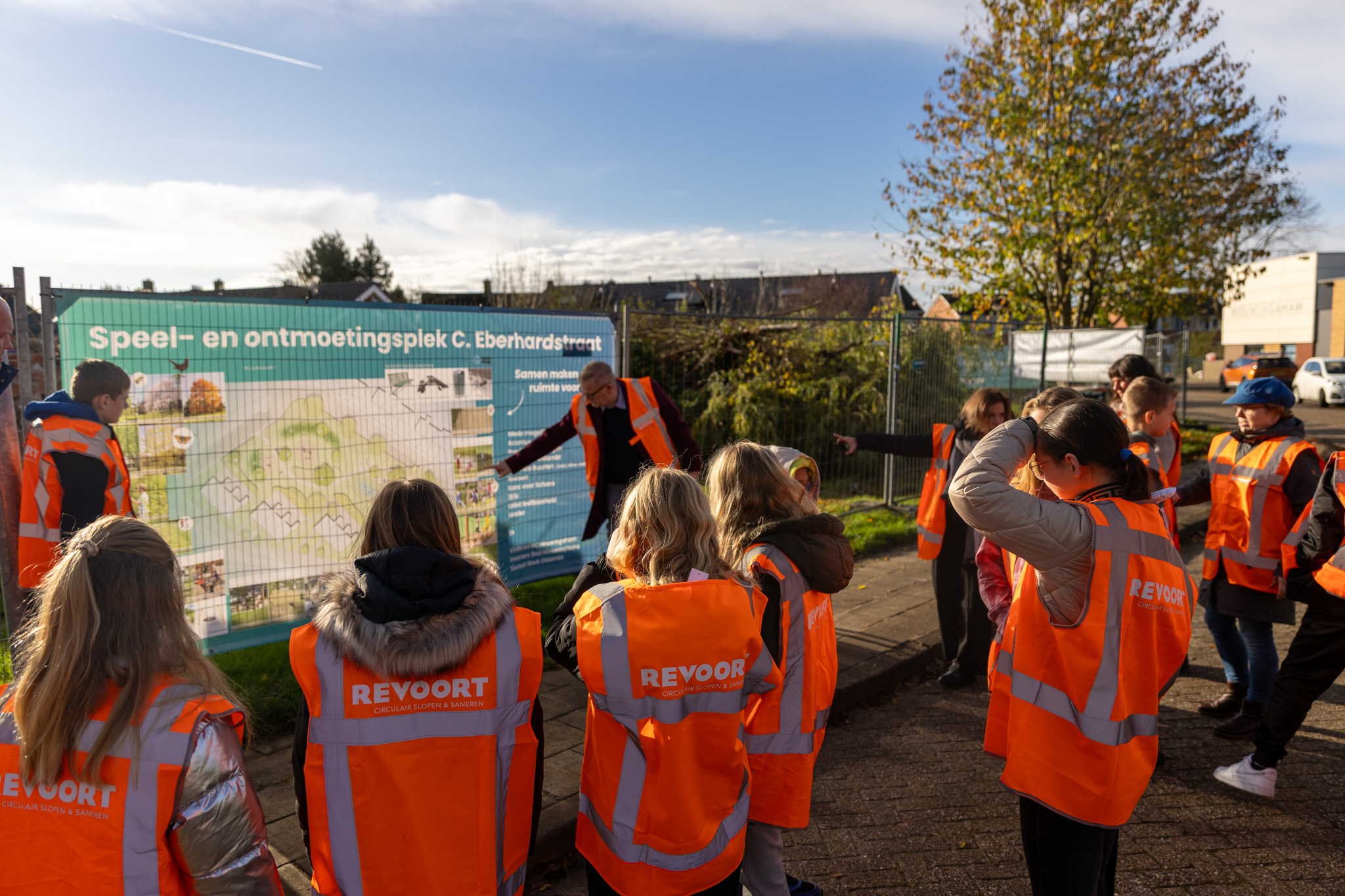 Groep kinderen in oranje hesjes bestuderen een bouwbord op een ontmoetingsplek onder begeleiding.