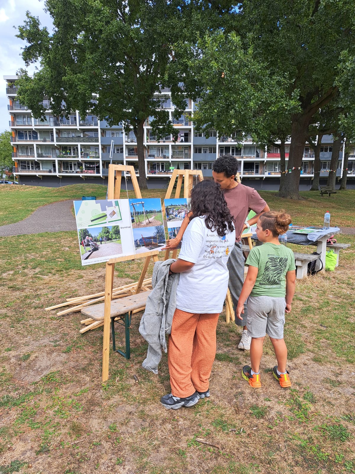 Drie mensen bekijken borden met foto's en informatie in een park met flats op de achtergrond.