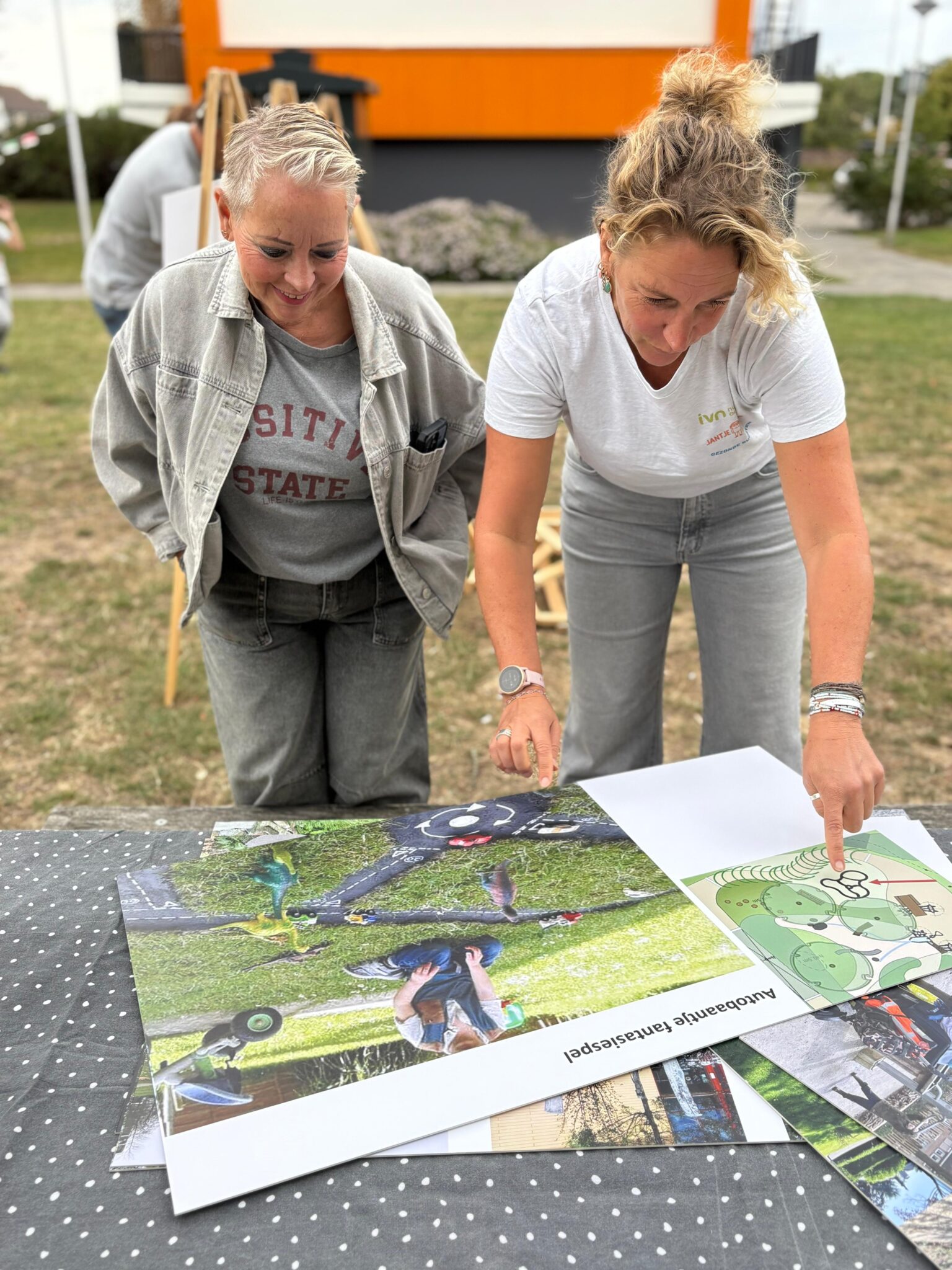 Twee vrouwen bekijken kaarten en foto's op een tafel in de buitenlucht.