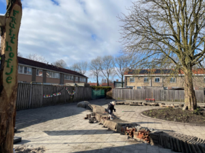 Speeltuin met houten bord "Fruithofje", kind spelend op een zonnige dag met kale bomen en huizen.