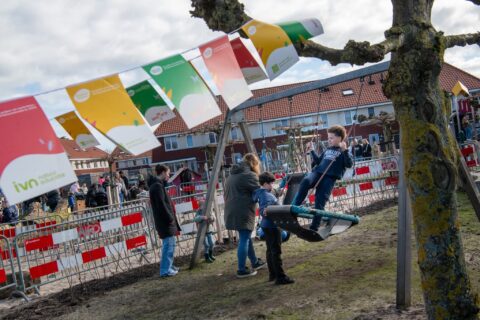 Kinderen spelen op een schommel in een speeltuin met kleurrijke vlaggen en toekijkende volwassenen.