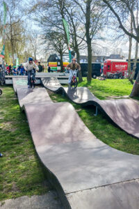 Twee kinderen fietsen op een golvende baan in een park met bomen en een rode foodtruck op de achtergrond.