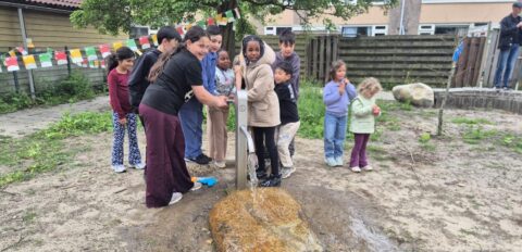 Groep kinderen speelt bij een openbare watertapkraan in een tuin met kleurrijke vlaggen erboven.