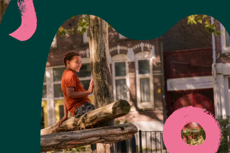 Een kind in een oranje shirt zit lachend op een boomstam, met een stadsgebouw op de achtergrond.