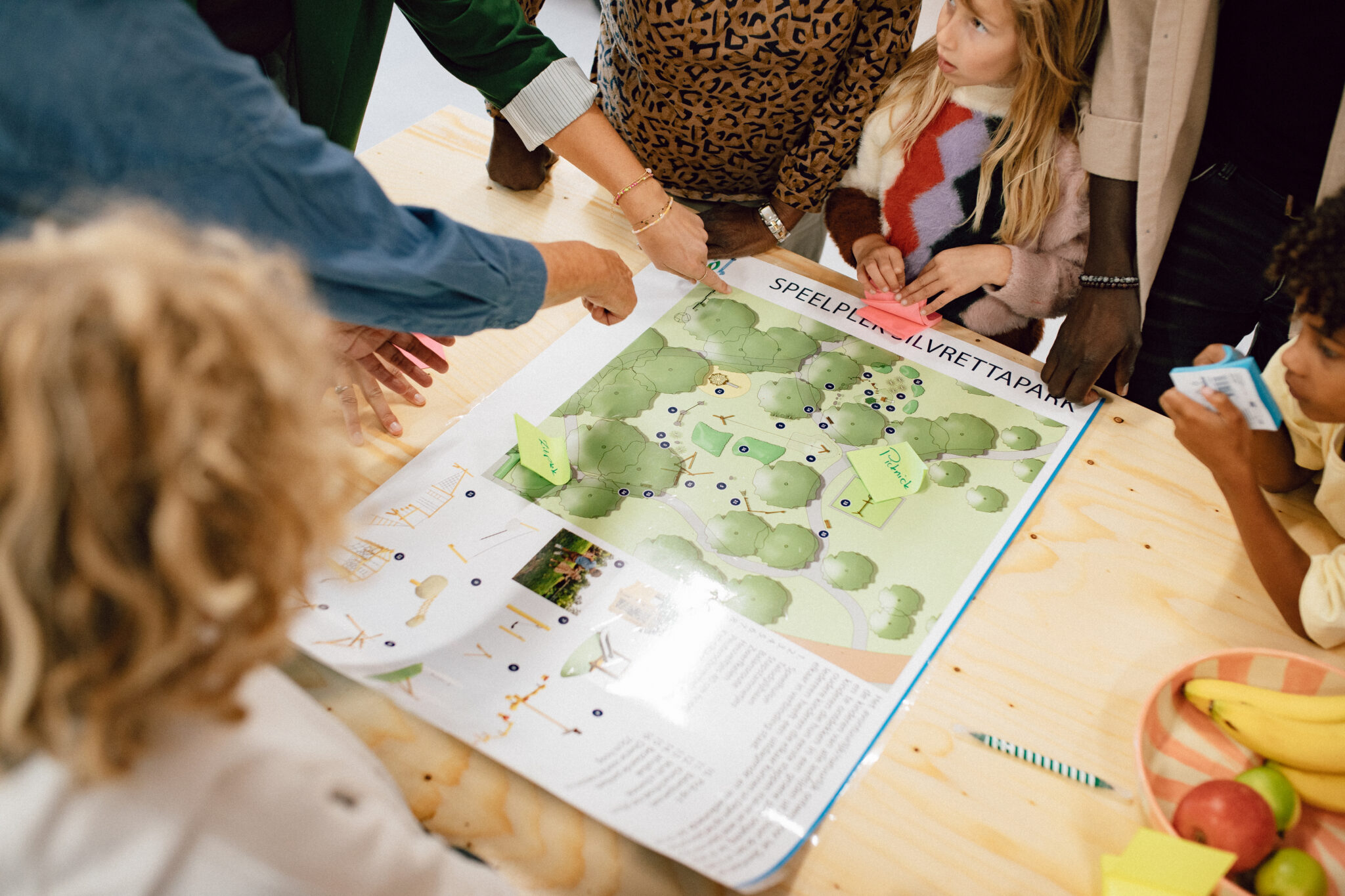 Een groep mensen bespreekt een speelplattegrond op een tafel. Kinderen kijken toe.