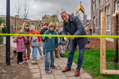 Een man en kind knippen een lint door voor een buitenspeelplaats, omringd door blije kinderen.