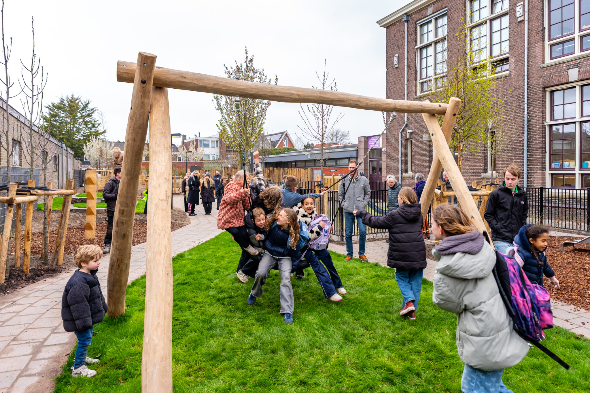 Kinderen spelen op een schommel in een schoolplein, omringd door volwassenen en een gebouw.