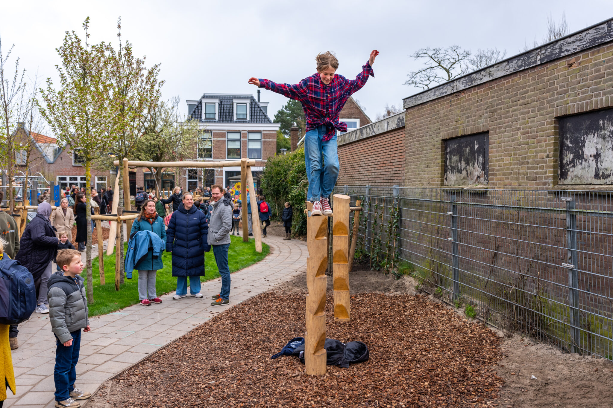 Kind balanceert op houten stapel in speeltuin, omringd door toekijkende mensen.