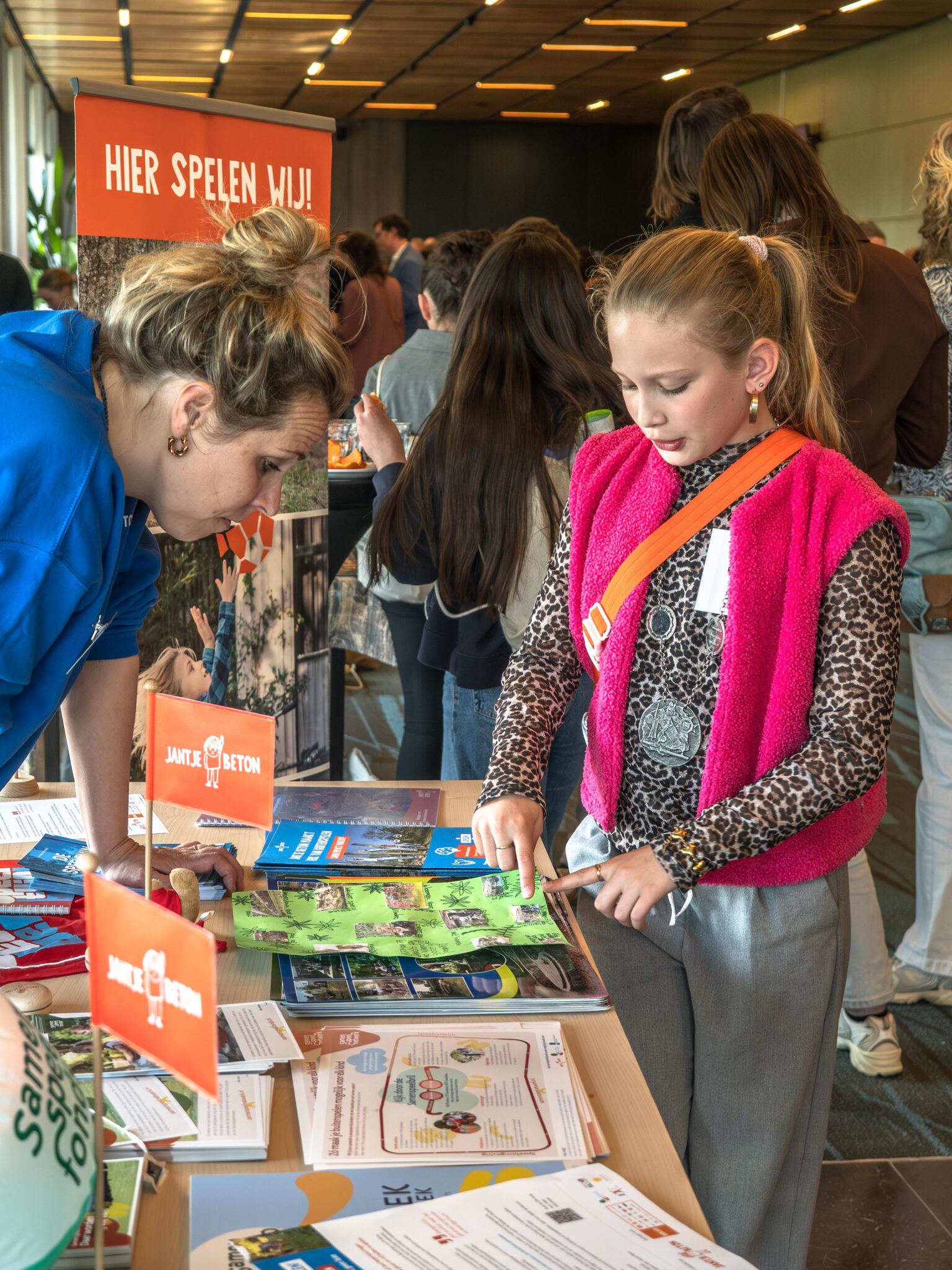 Meisje kijkt naar folders op een stand met 'Hier spelen wij!' poster op achtergrond.