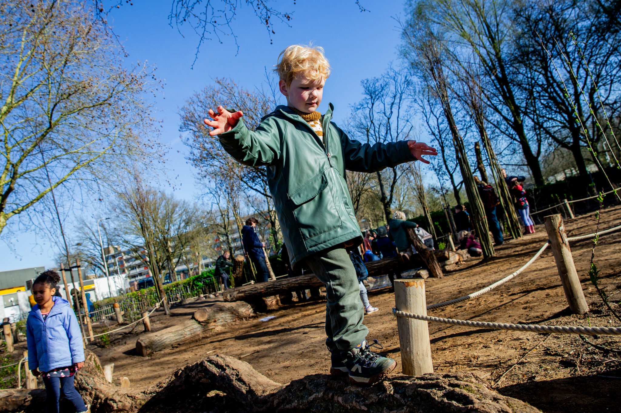 Kinderen spelen in een speeltuin, jongen balanceert op een boomstam, zonovergoten omgeving.