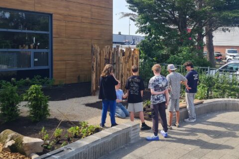 Groep tieners staat bij houten kunstwerk naast modern gebouw in een groene omgeving.