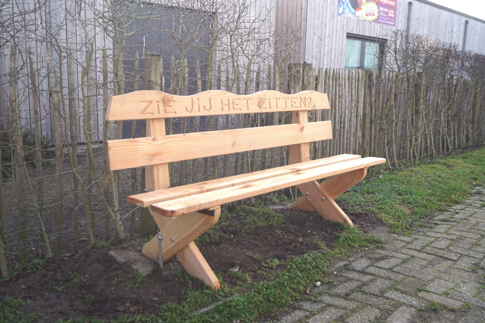 Houten bankje met tekst "Zie jij het zitten?" naast een pad, omringd door heg en gebouw.