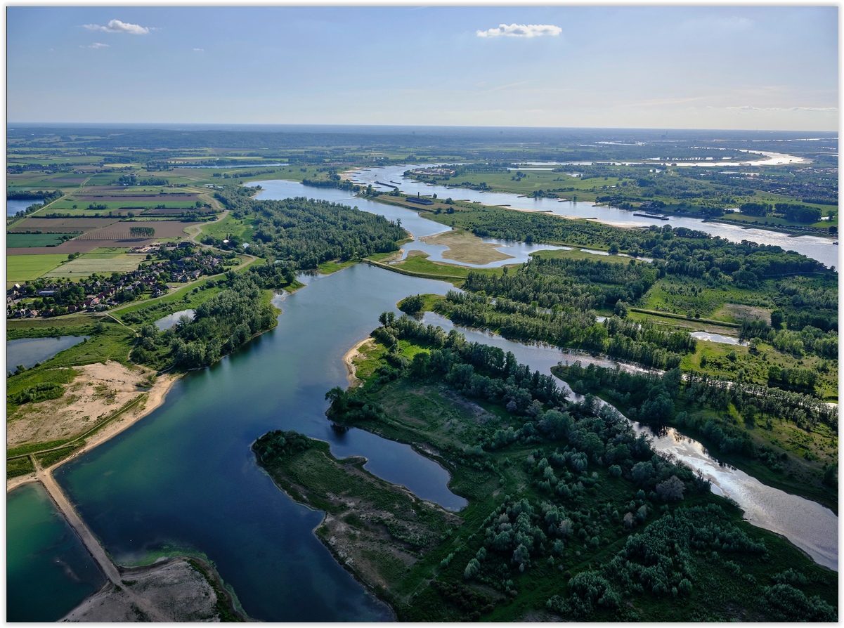 Luchtfoto van een rivierlandschap met groene velden en bosrijke gebieden onder een blauwe hemel.