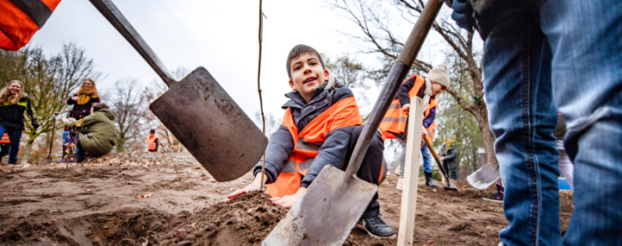 Kinderen planten bomen in een bos met schoppen, een jongen in een oranje hes kijkt omhoog.