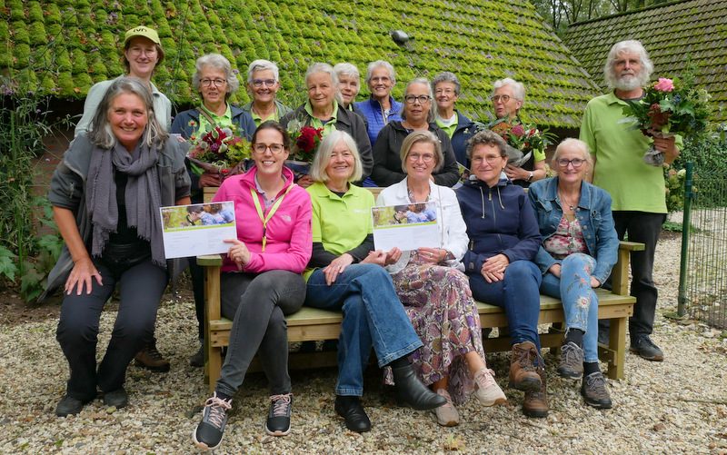 Groep mensen, sommigen met bloemen, poseert buiten bij met mos begroeide daken. Twee houden certificaten vast.