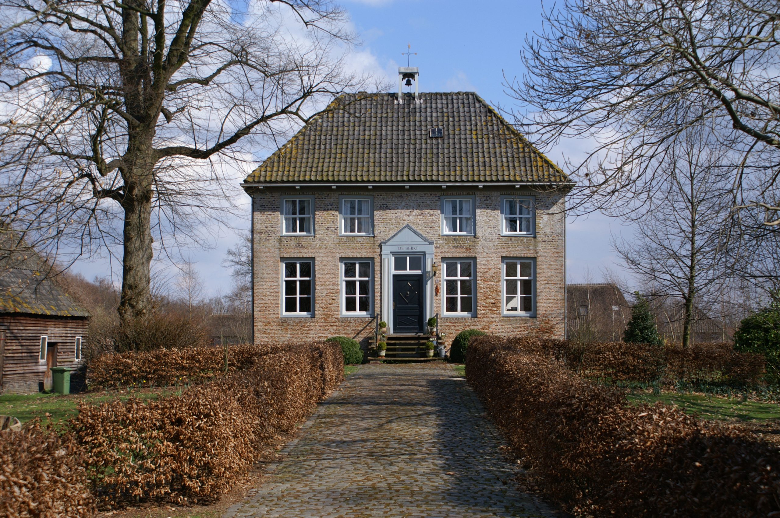 Bakstenen huis met blauwe deur en ramen, omringd door kale bomen en een heg, onder een blauwe lucht.