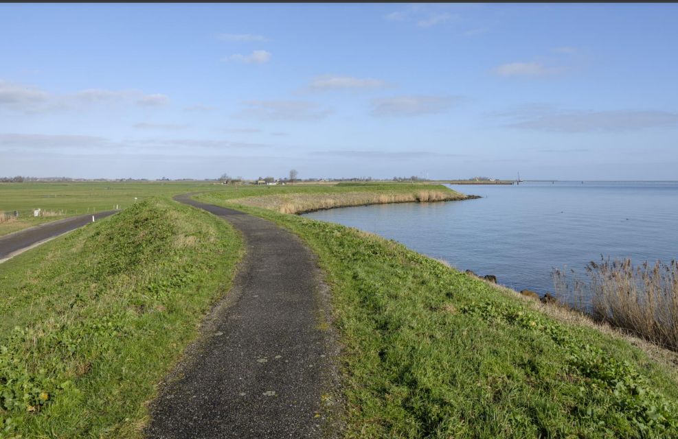 Gewelfde dijkweg langs een kalm meer onder een blauwe hemel met verspreide wolken.