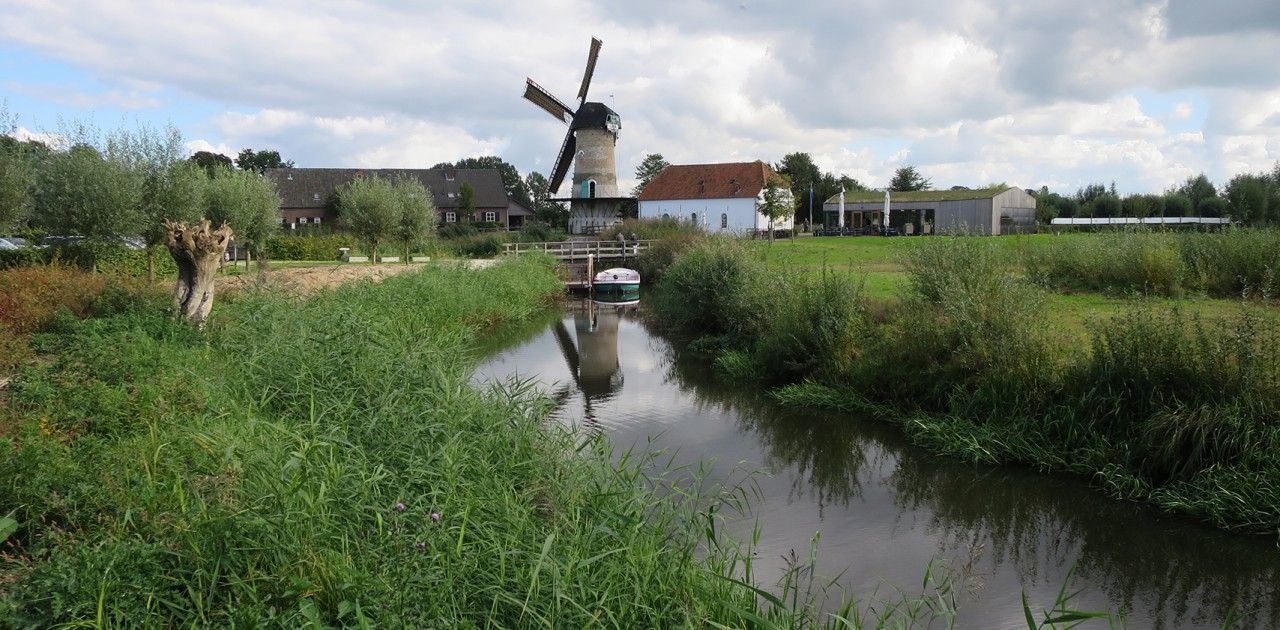 Windmolen bij een rivier met omringend groen, gebouwen en bewolkte lucht.